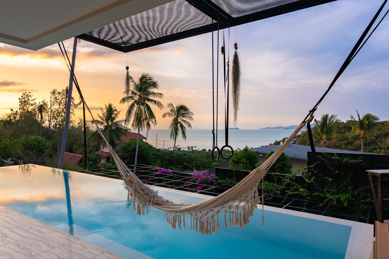 A hammock over the pool at golden hour, palm trees and sea behind